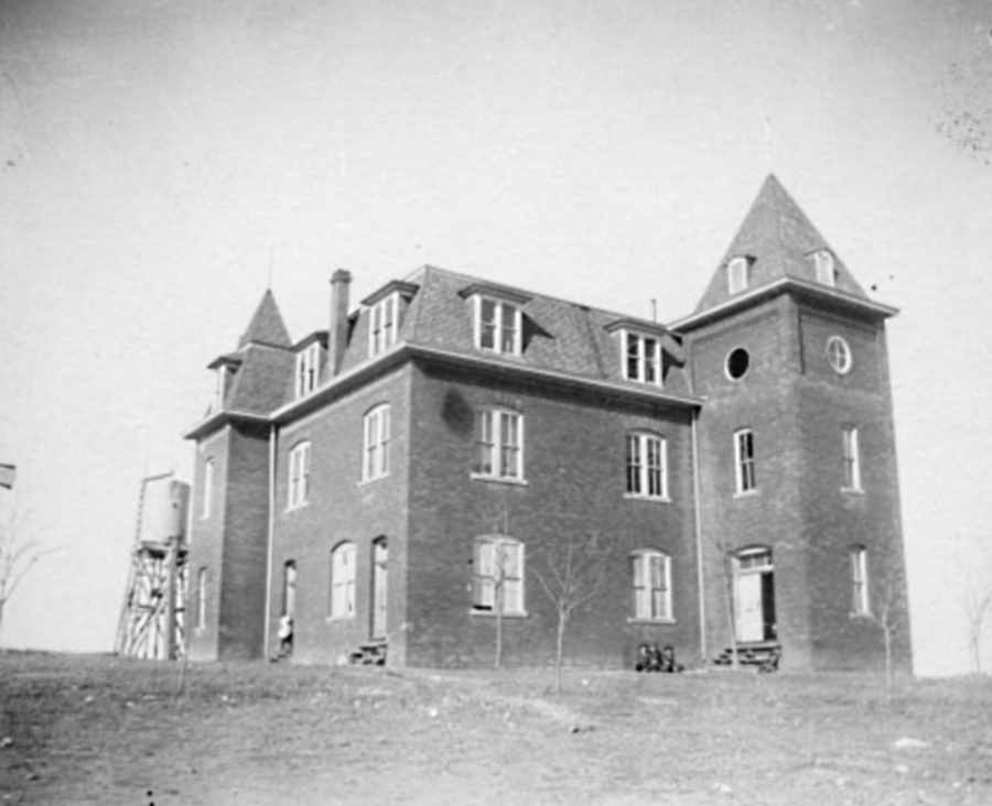 Photograph of the main building of Clarendon College in Clarendon, Texas in 1906. There are three children sitting on the ground in front of the building, and a girl is about to enter one of the doors. 